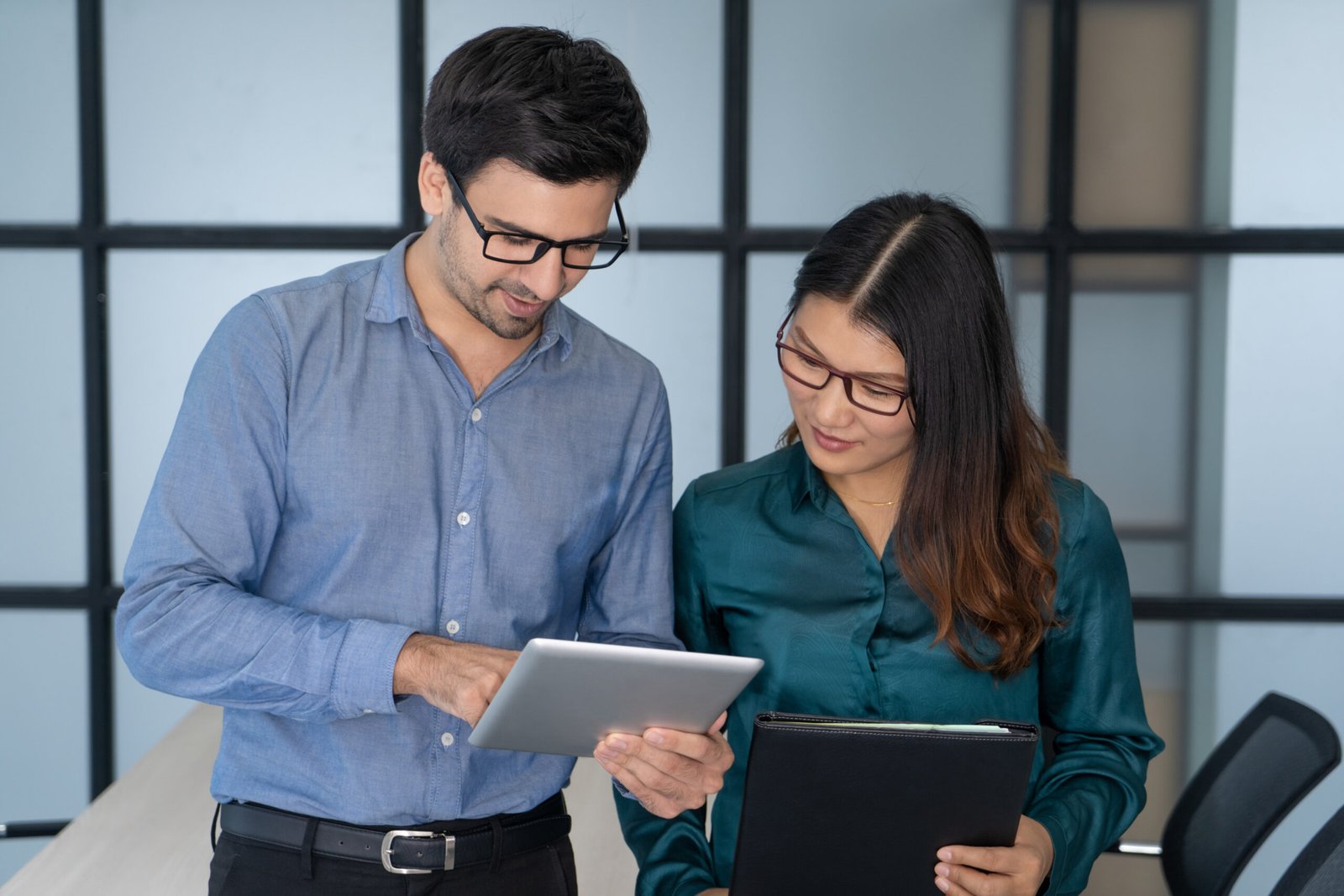 Caucasian supervisor helping new Asian employee. Friendly office worker showing to young colleague project details on tablet screen. Training and corporate help concept